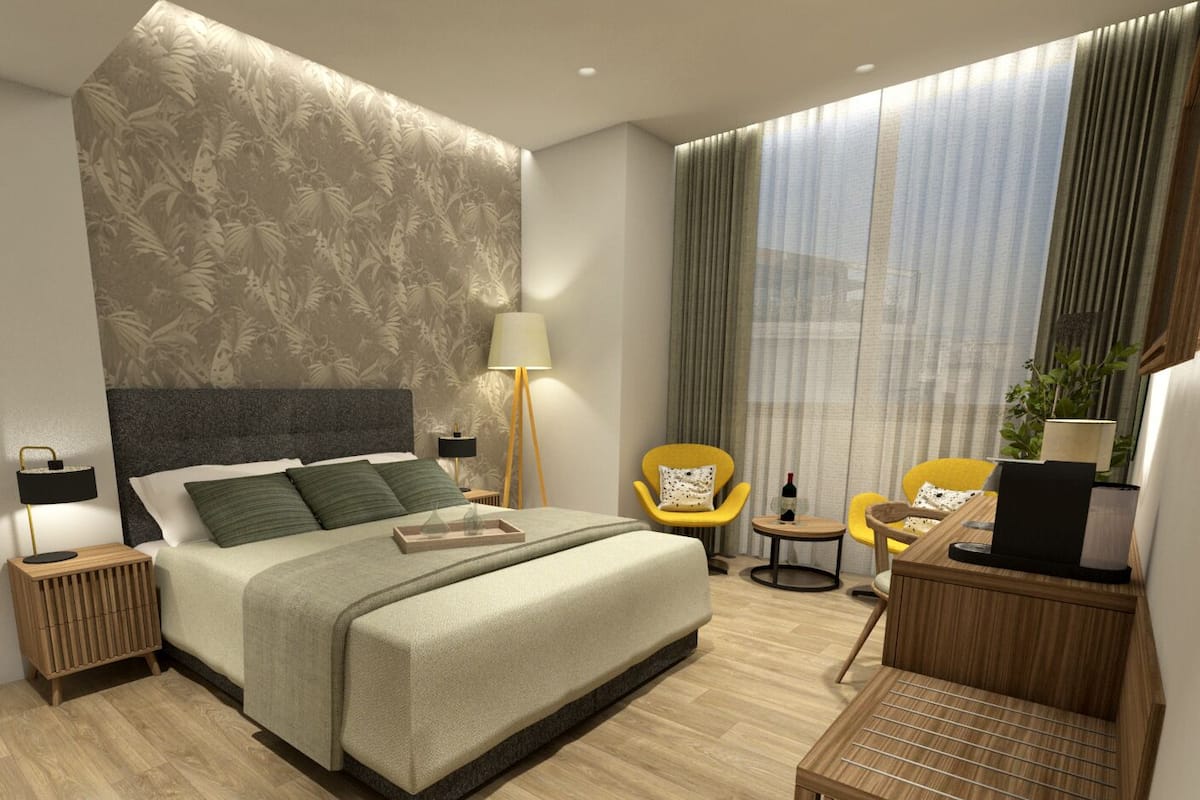 A modern hotel room with a large bed, a desk, two chairs, a lamp, and a view of the city through large windows.