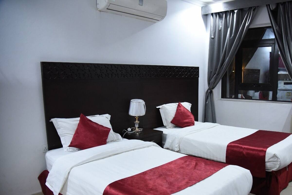 A hotel room with two beds, white bedding, red accents, a dark headboard, a bedside lamp, and a window with curtains.