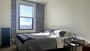 Une chambre à coucher avec un lit, un radiateur, une fenêtre avec des stores et une petite table avec une lampe.
