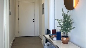 Un couloir avec une porte blanche au fond, un miroir fixé au mur, une plante en pot et une poubelle.
