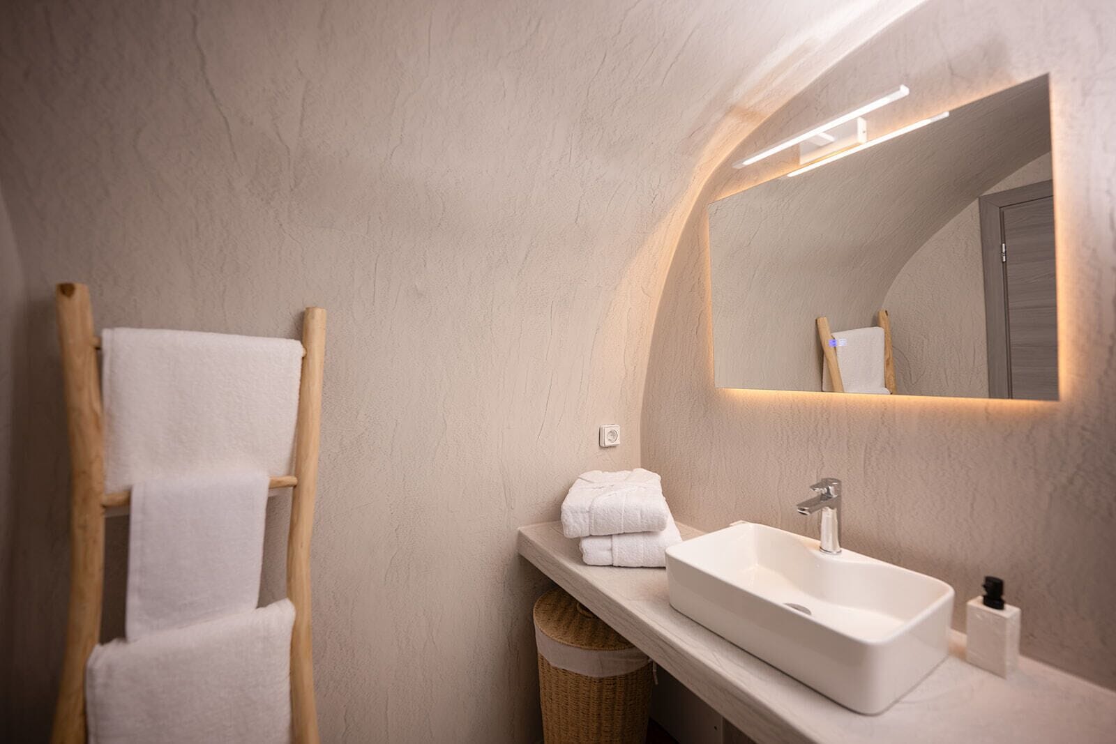 A modern bathroom with a wooden towel rack, a white sink, and a mirror with integrated lighting.