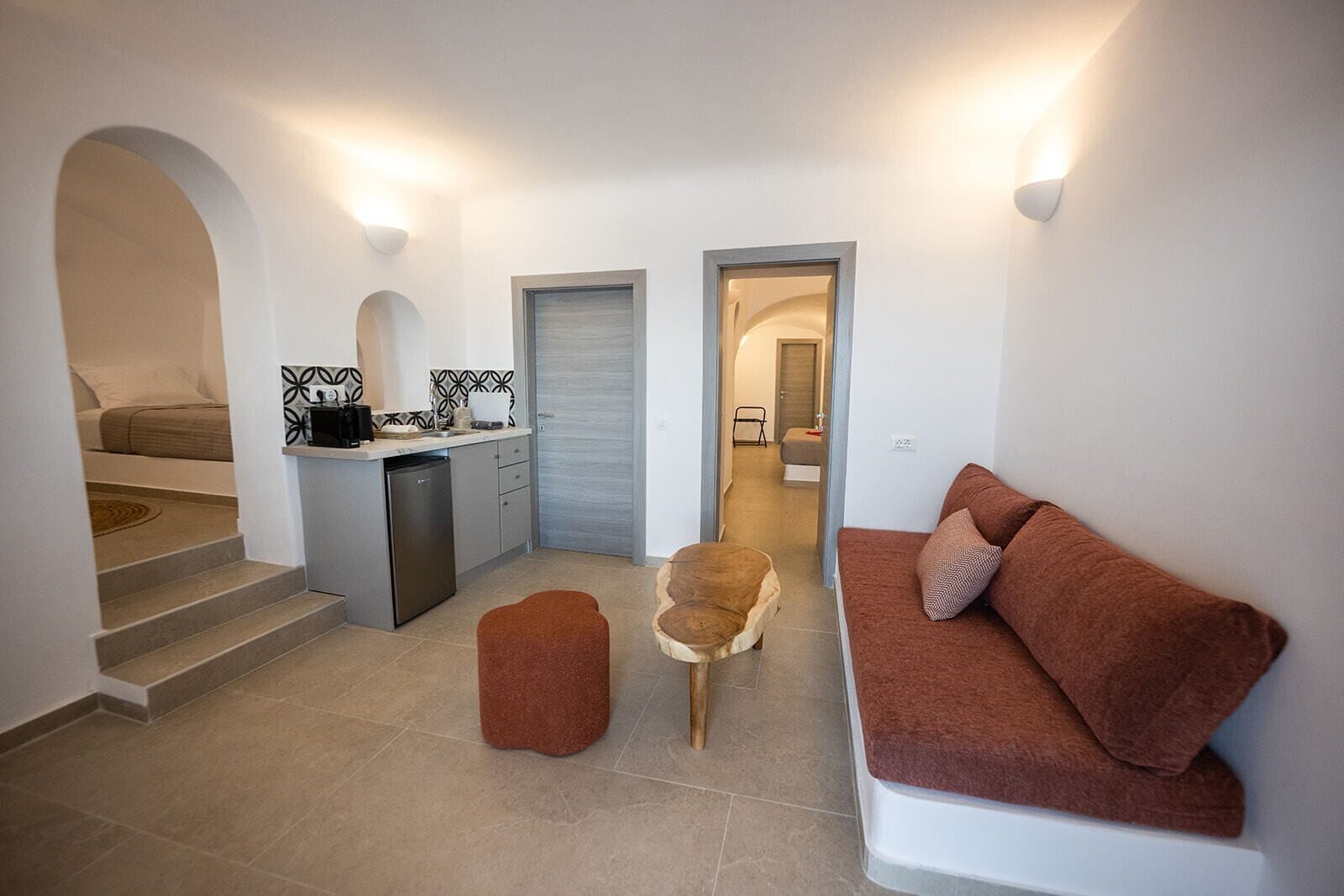 A compact living space with a kitchenette, a sofa, a wooden stool, and a staircase leading to a bedroom.