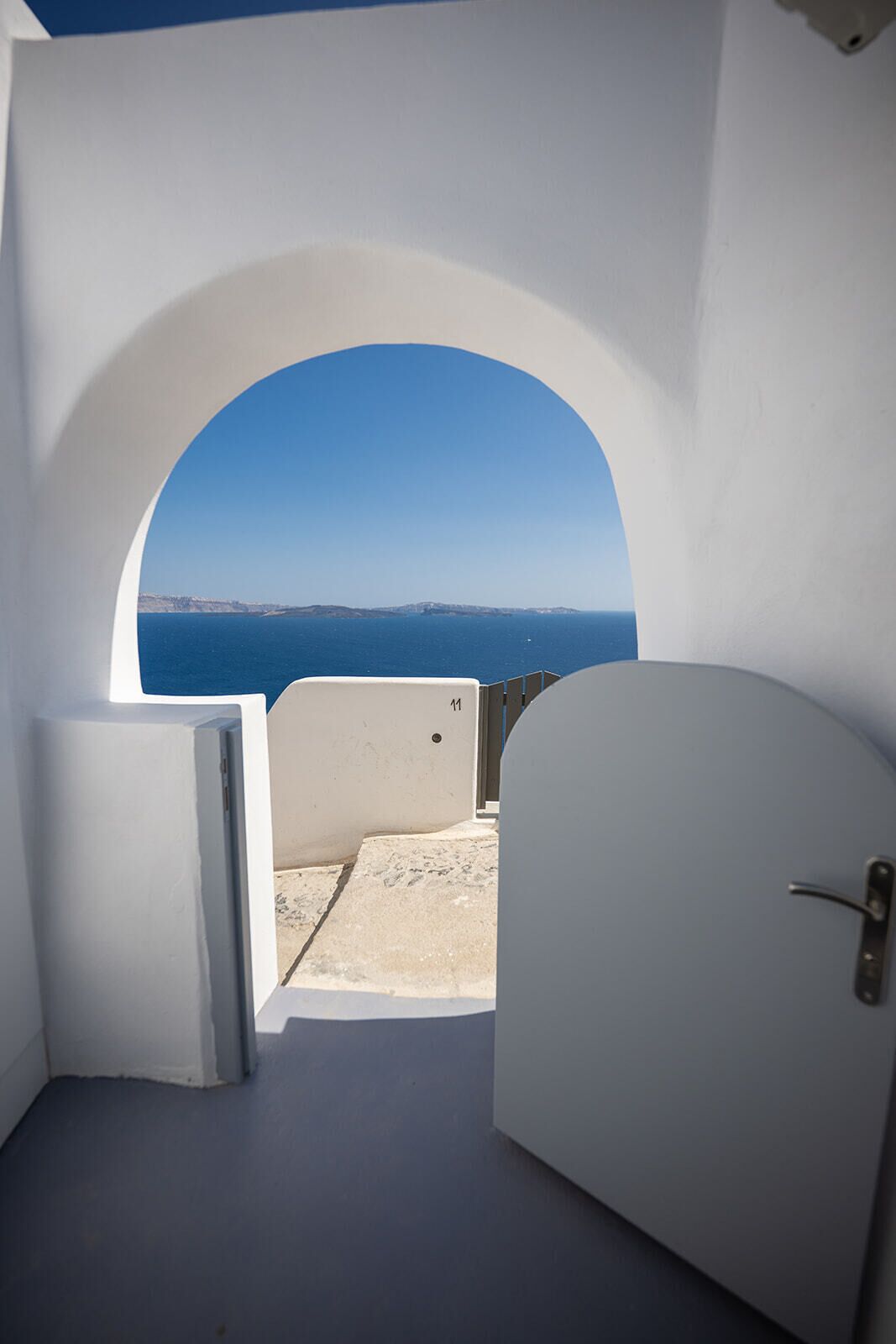 Une pièce avec vue sur la mer, donnant sur celle-ci par une fenêtre en arc de cercle, une porte et un petit espace extérieur doté d’un sol en béton.