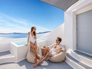 Una pareja en la terraza de una piscina infinita blanca, con vistas claras al mar y a la costa distante.