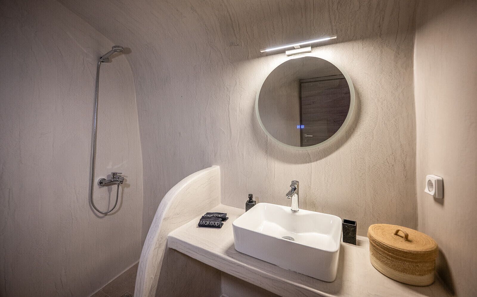 A bathroom with a white sink, a handheld showerhead, a round mirror, and a small wooden container.