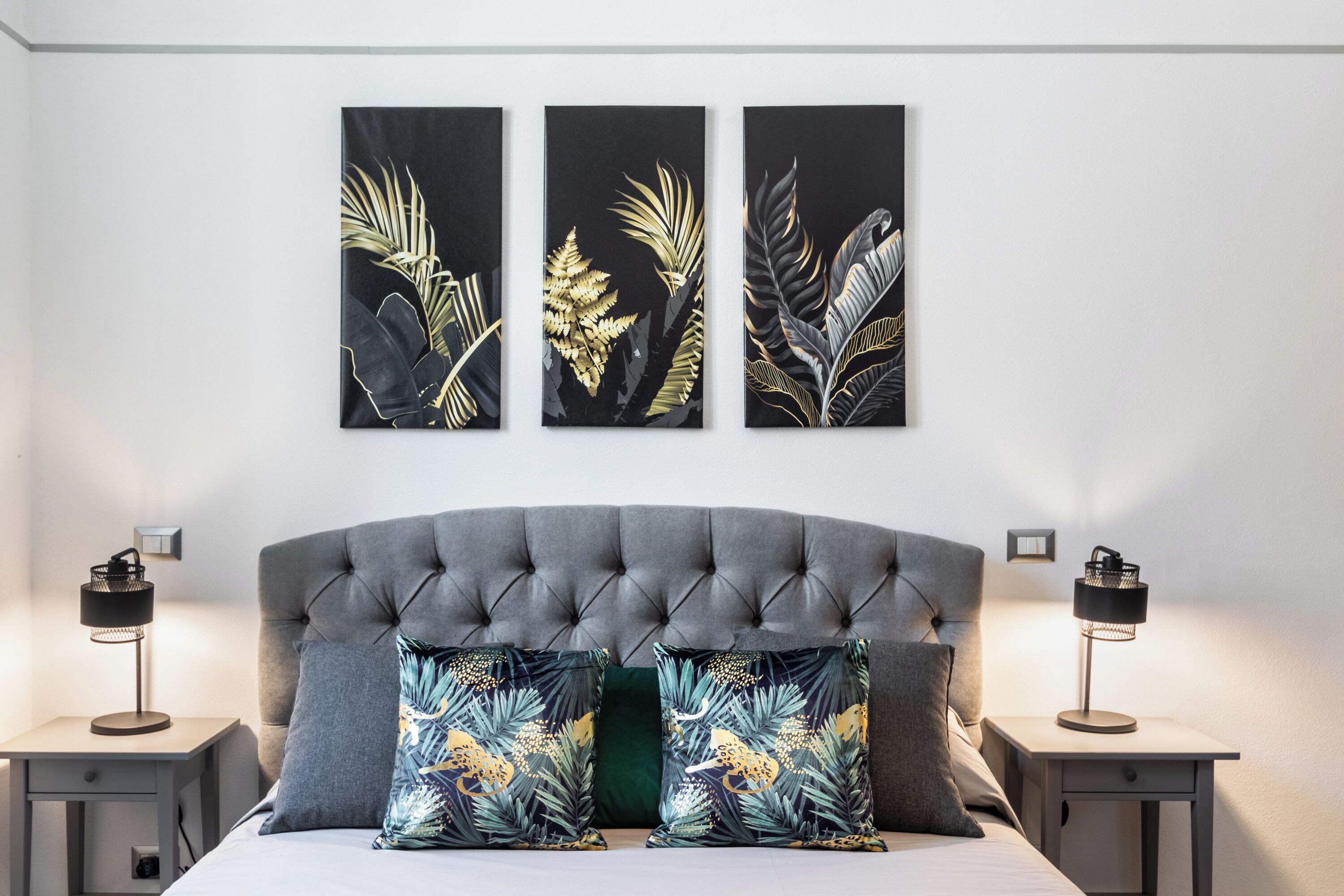 Een slaapkamer met een gestoffeerd hoofdeinde, twee nachtkastjes met lampen en drie wanddecoraties.