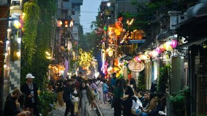 A bustling street scene with people walking and sitting on a narrow path, surrounded by buildings with traditional architecture and colorful lanterns. - Homestay-Kim Mã -Giang Văn Minh (Hanoi)