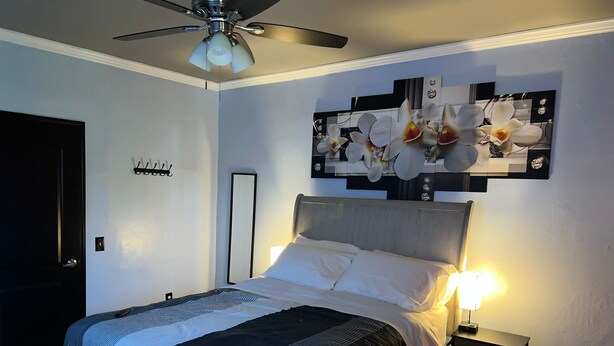 Un dormitorio con una cama, una mesita de noche, un ventilador de techo y una obra de arte en la pared con orquídeas.