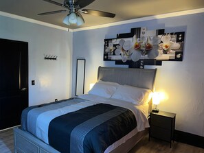 Een slaapkamer met een bed, een nachtkastje, een plafondventilator en een kunstwerk van orchideeën aan de muur.
