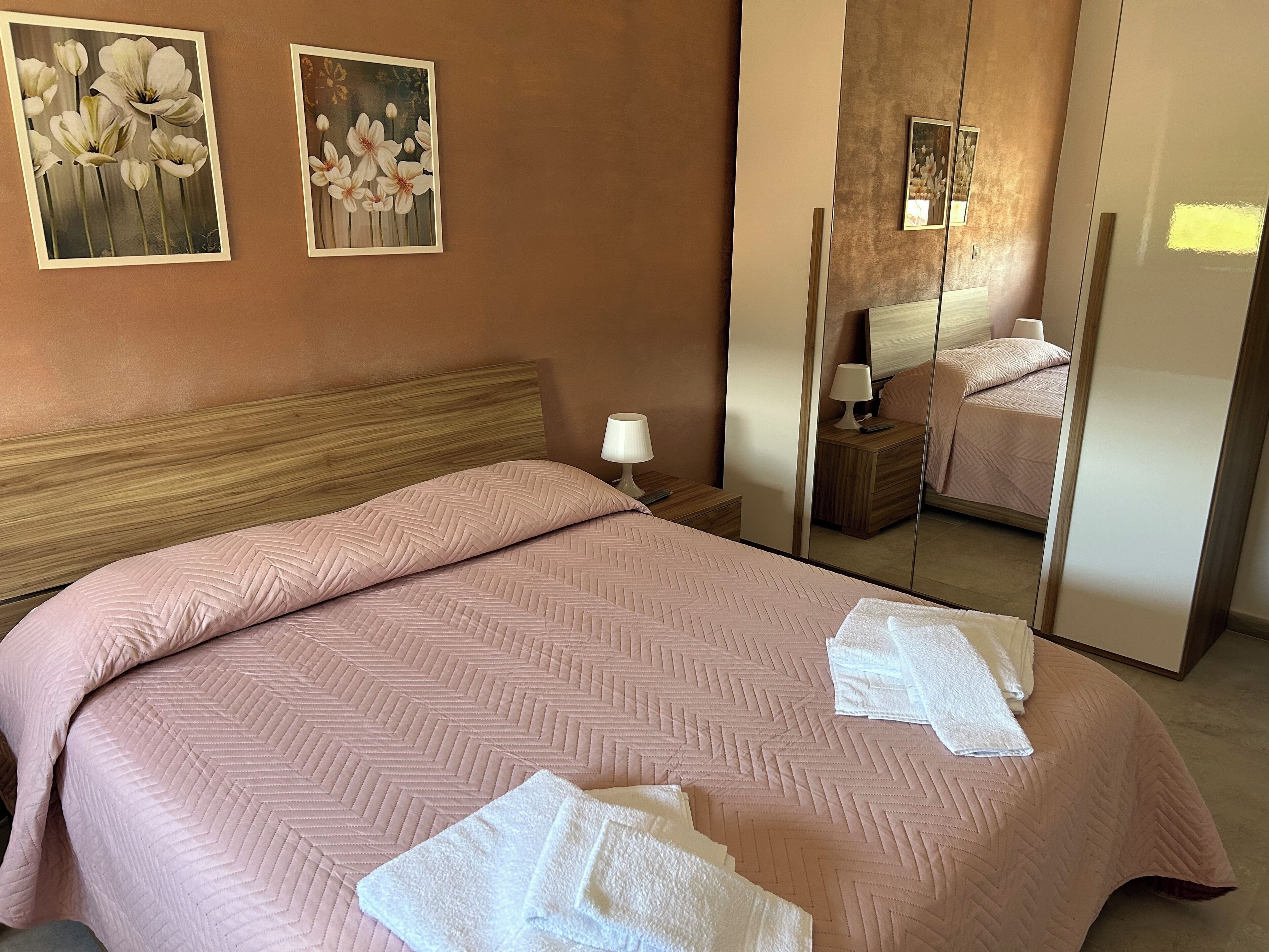 Una camera d'albergo con un letto grande, una testiera in legno, un comodino con una lampada e uno specchio a figura intera.