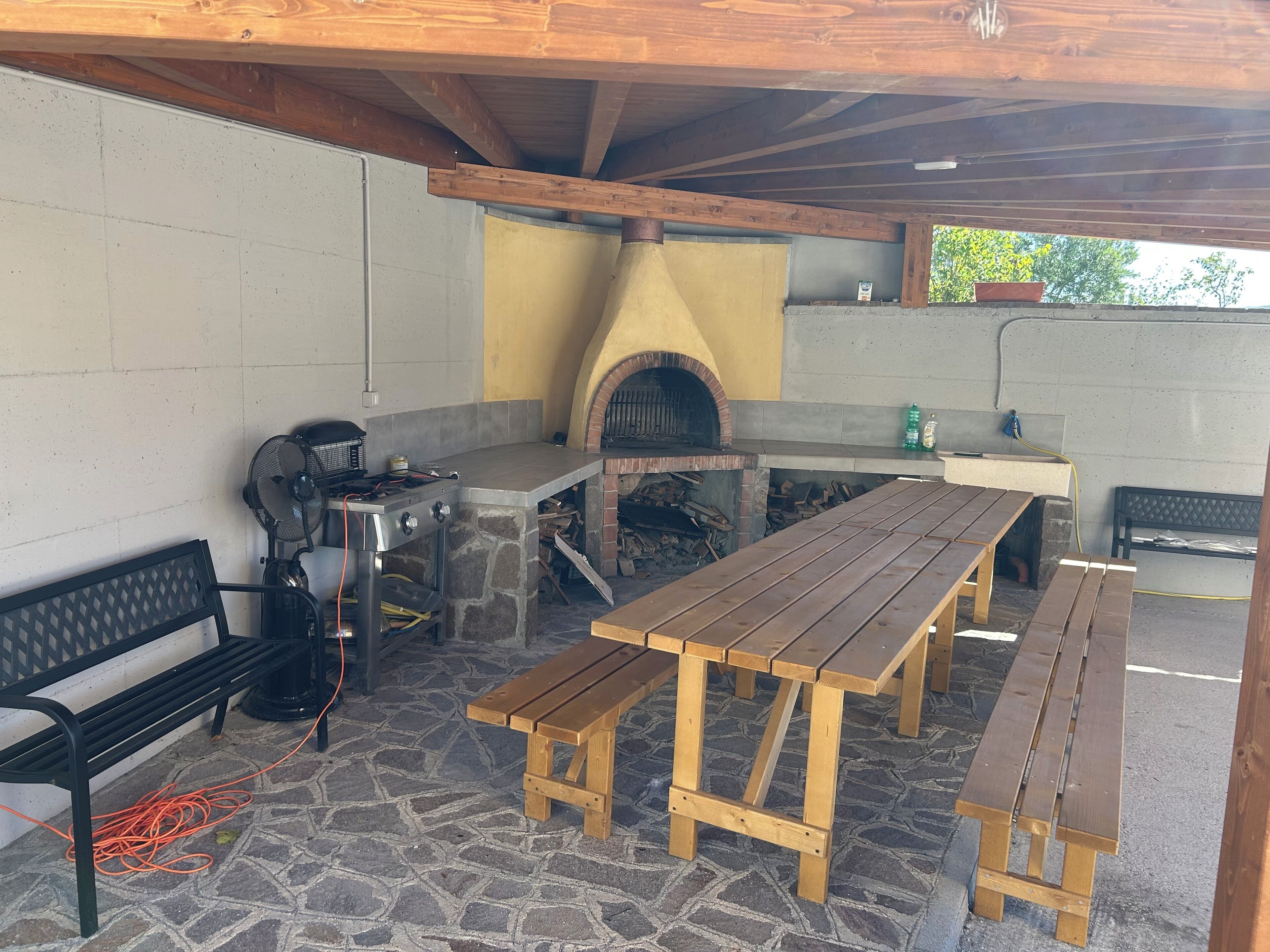 An outdoor patio with a stone floor, wooden benches, a grill, and a brick oven.