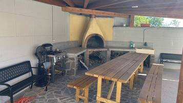 An outdoor patio with a stone floor, wooden benches, a grill, and a brick oven.