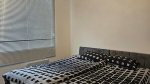 A neatly made bed with a checkered bedspread and two pillows, a radiator below the window, and a plain wall.