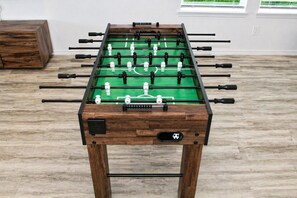 A wooden foosball table with a green playing surface, set on a wooden floor in a room with white walls and a window. - Modern 3 BR New House with Grill and Games (Pattison)