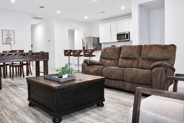 A modern living room with a brown sofa, a wooden coffee table, and a dining area with a wooden table and chairs. - Modern 3 BR New House with Grill and Games (Pattison)