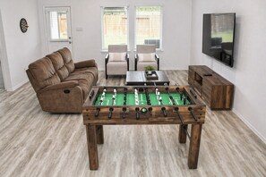 Un salon avec un baby-foot, un canapé marron, deux fauteuils, une table basse et un téléviseur fixé au mur.