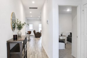 A hallway with a mirror, a plant, a desk, a chair, and a bed in the adjacent room.