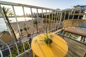 A balcony with a yellow table and a wicker chair, overlooking a beach with palm trees and a cityscape in the background. - Brisas del Poniente-Deluxe Room Ocean View (Benidorm)