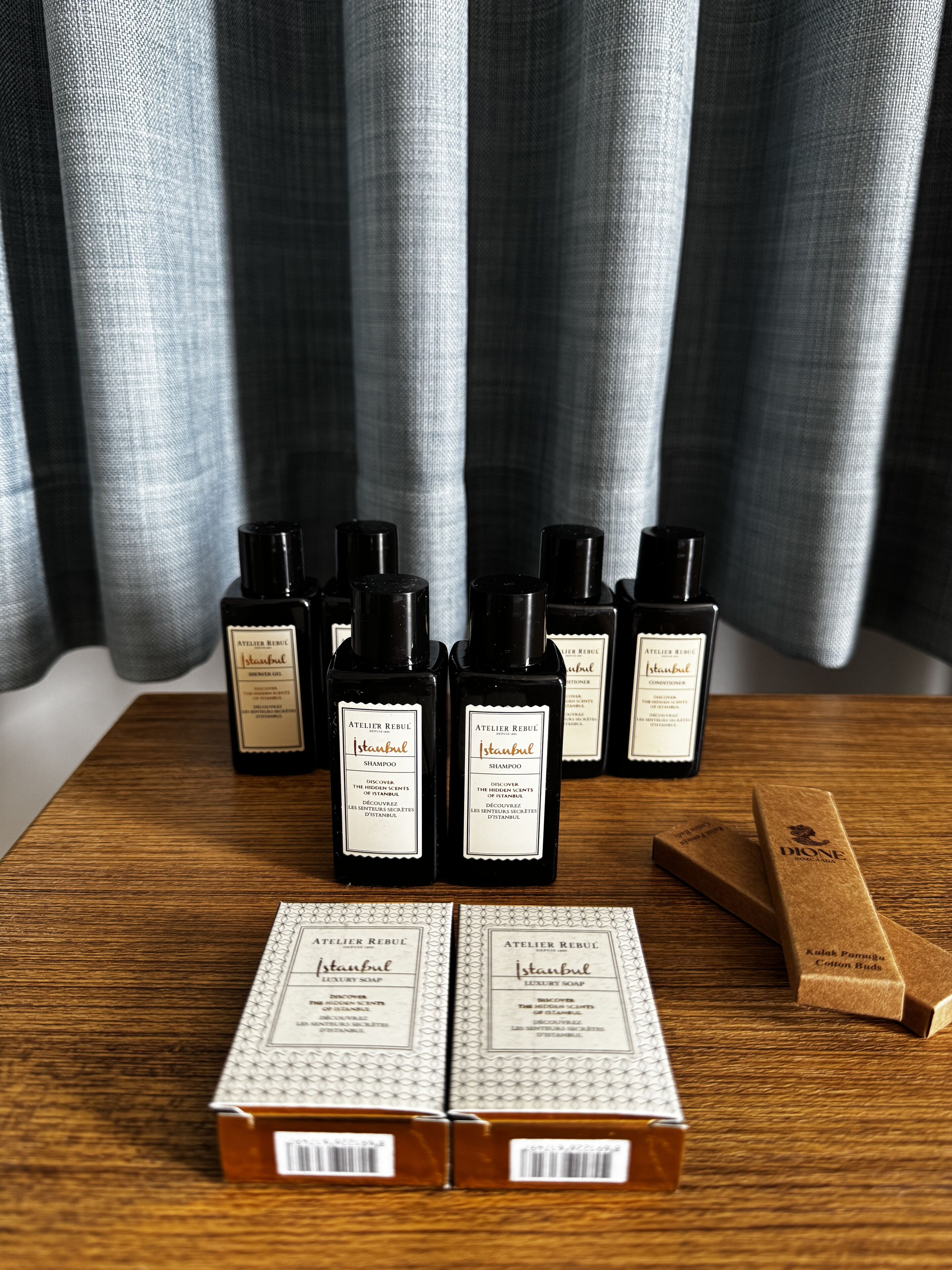 Una mesa de madera con varias botellas de productos con motivos de Estambul, incluyendo champú y jabón de lujo, y dos cajas de bastoncillos de algodón.