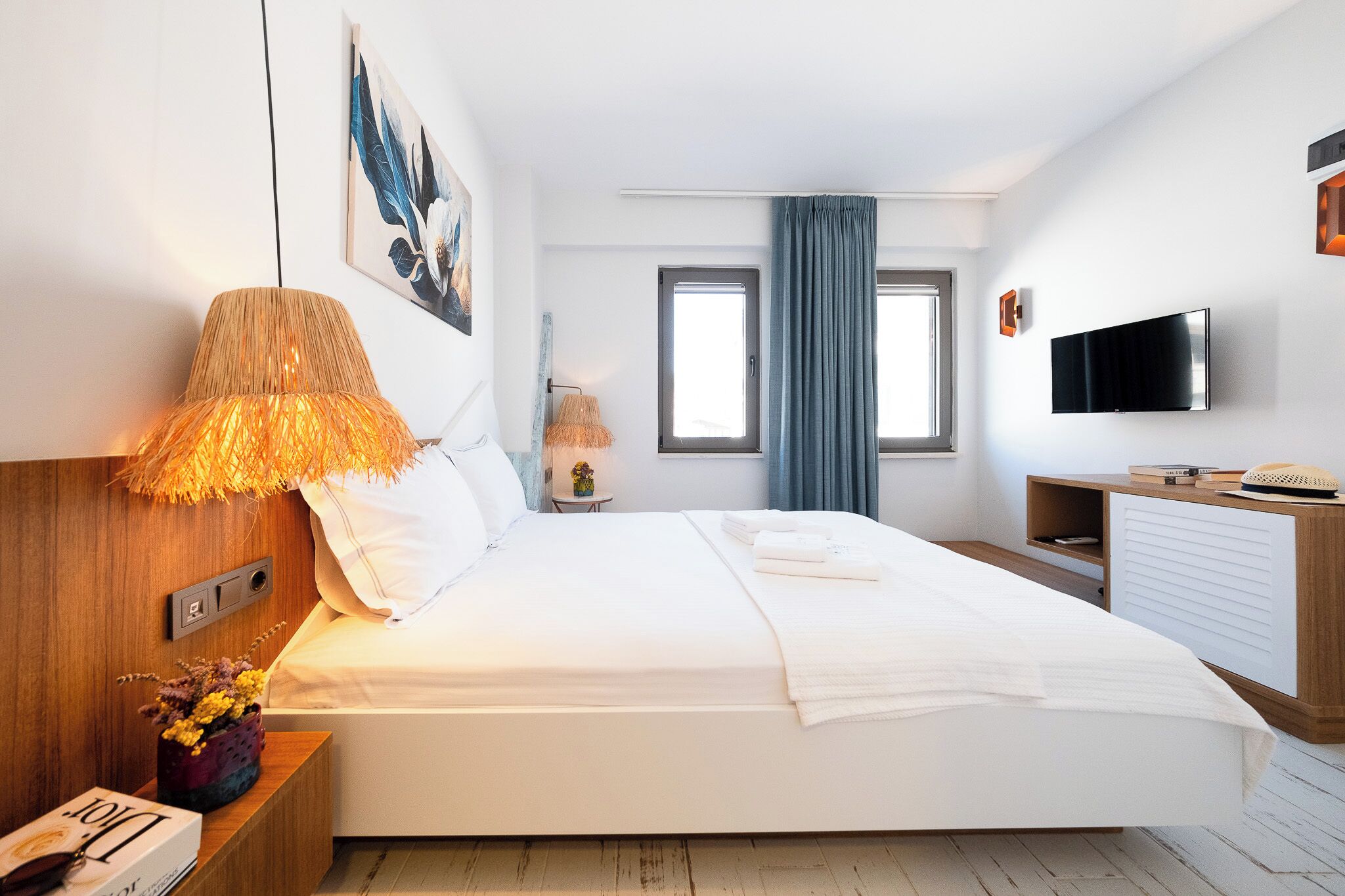 A modern hotel room with a large bed, a flat-screen TV, a wooden nightstand, and a window with blue curtains.