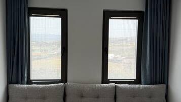 Una habitación moderna con sofá gris, dos ventanas con cortinas y vistas a un paisaje urbano.