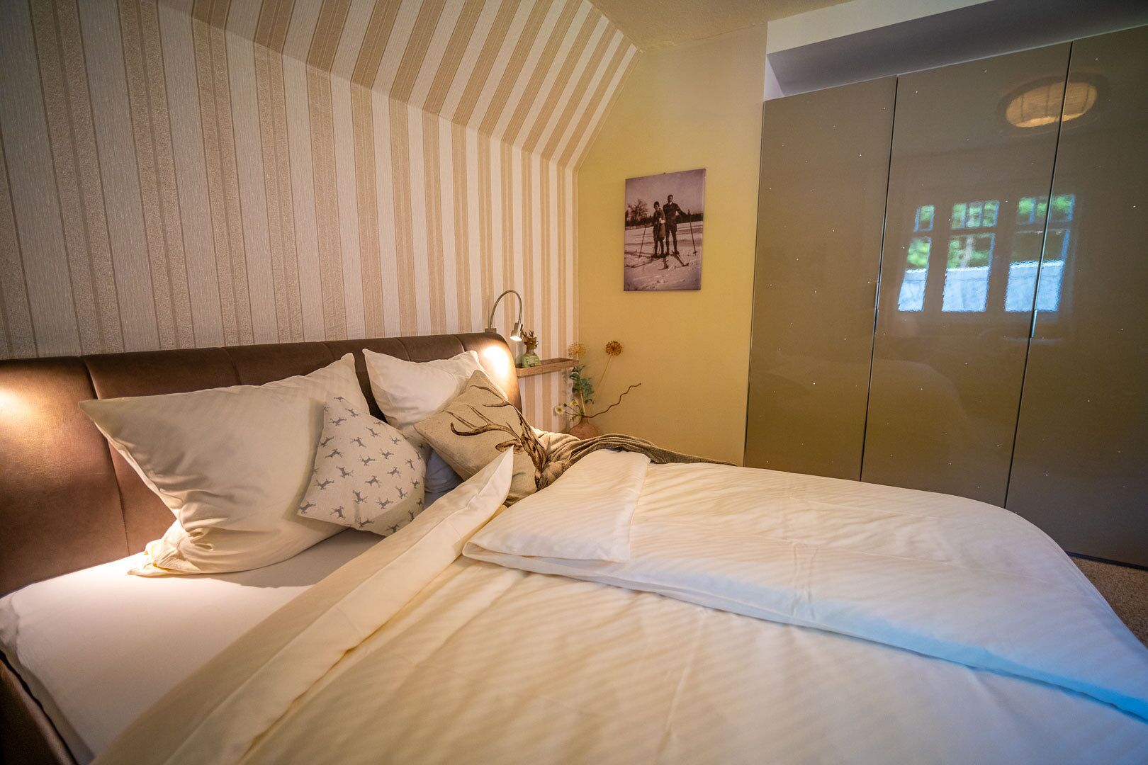 Un lit bien fait, avec du linge de lit blanc et une variété d’oreillers, une lampe de chevet et une photographie encadrée accrochée au mur.