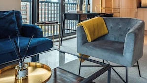 Una sala moderna con sofá, sillón y mesa de centro, ventanas amplias con vistas a la ciudad, y una zona de cocina con armarios y una lámpara colgante.