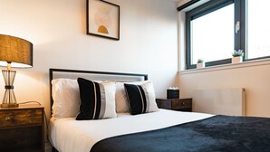 Un dormitorio ordenado con una cama, una mesita de noche, una lámpara, un cuadro en la pared y una ventana con vistas.