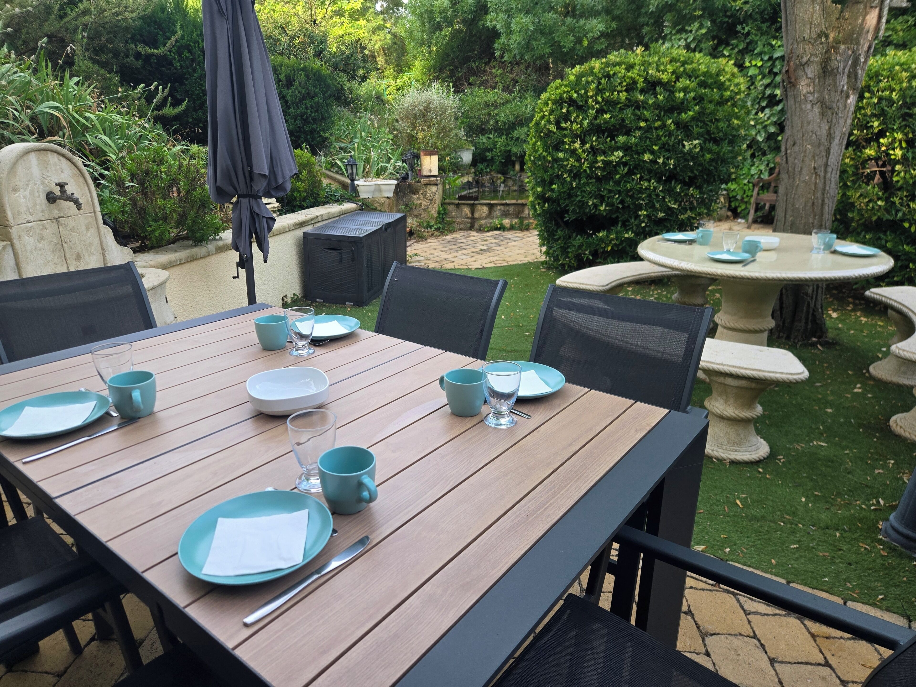 Une table en bois, dressée avec des assiettes, des tasses et des couverts, se trouve dans un jardin agrémenté d’une fontaine en pierre et de verdure.