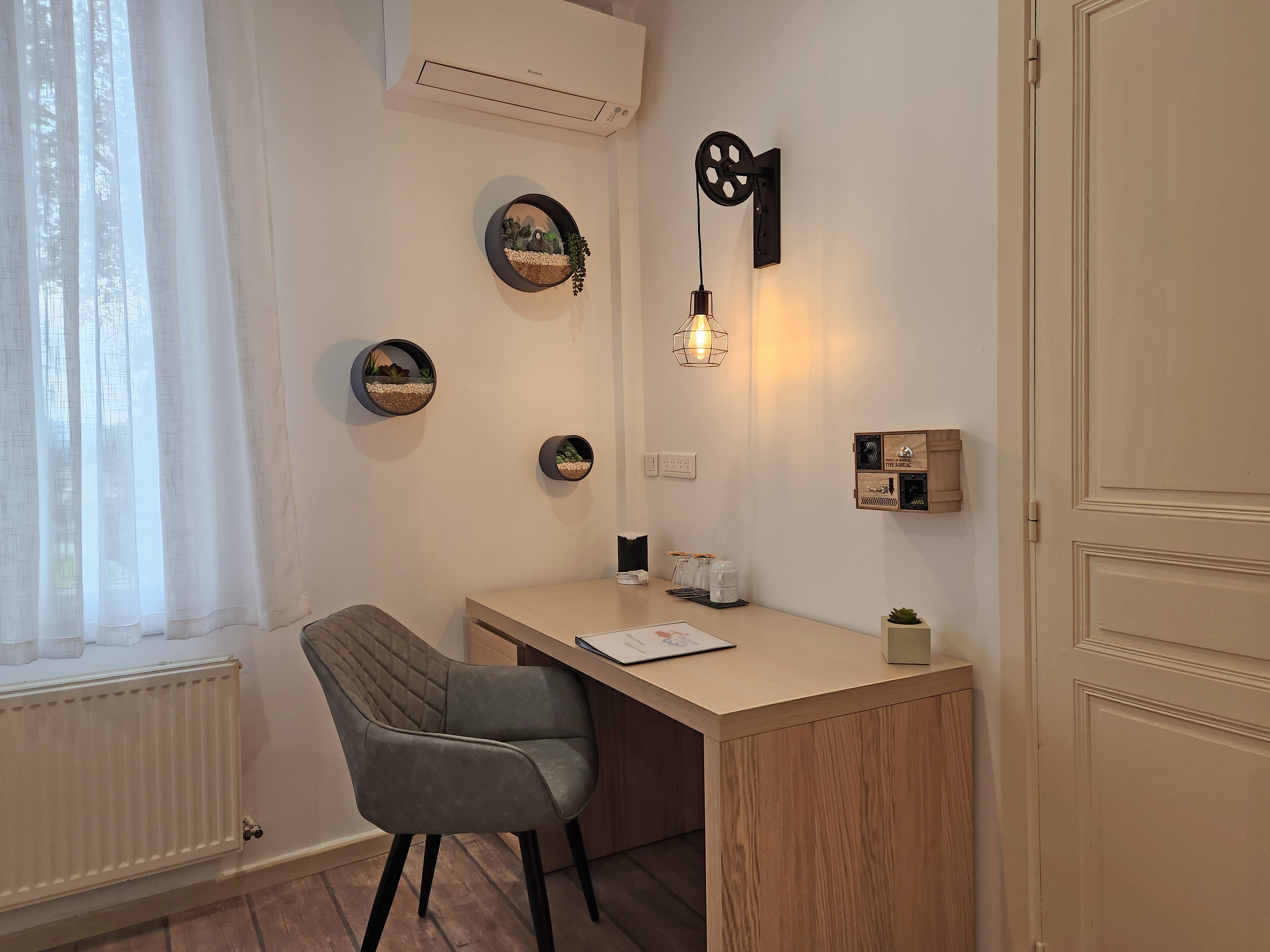 Una habitación pequeña y bien iluminada con un escritorio, una silla y varios adornos en las paredes, incluyendo cuadros enmarcados y una lámpara fijada a la pared.