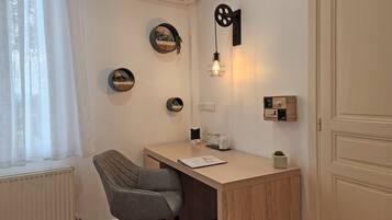 Una habitación pequeña y bien iluminada con un escritorio, una silla y varios adornos en las paredes, incluyendo cuadros enmarcados y una lámpara fijada a la pared.