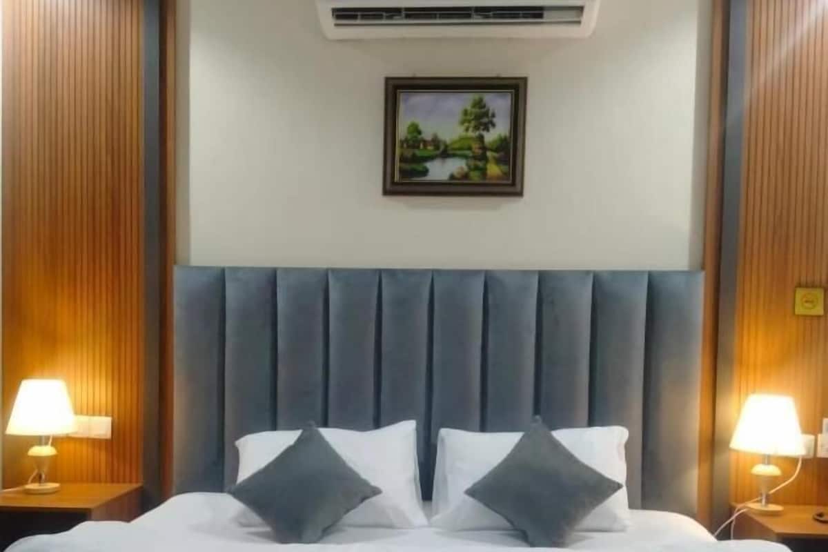 A hotel room with a large bed, two bedside lamps, an air conditioning unit, and a framed picture on the wall.