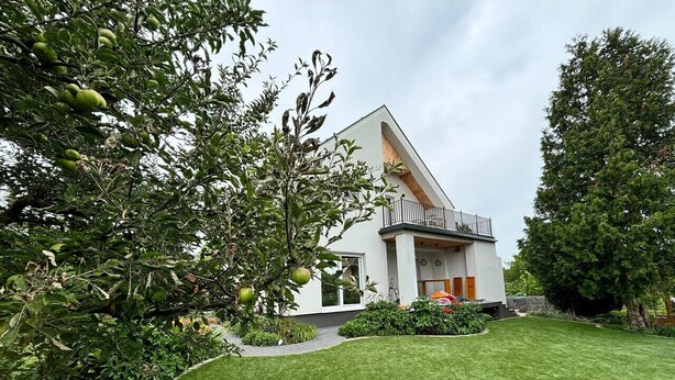 Una casa moderna con techo inclinado, césped cuidado y diversidad de árboles y arbustos.