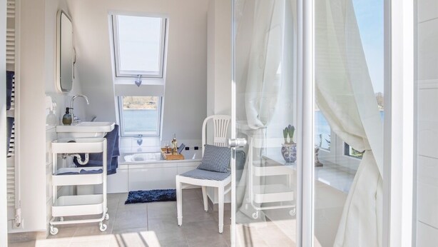 A modern bathroom with a white sink, a window, a chair, and a glass shower enclosure. - House Langsee 2 (Kosel)