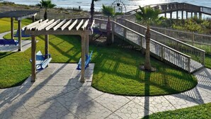 A coastal walkway with a wooden pergola, palm trees, and a view of the ocean. - Salty Sea Wishes - Ocean Views, Beach Access, Family Vibes (Galveston)