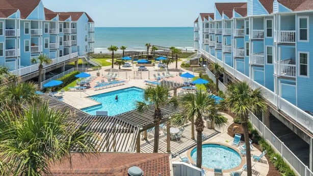 A resort with multiple blue buildings, a central swimming pool, and a beach view. - Salty Sea Wishes - Ocean Views, Beach Access, Family Vibes (Galveston)
