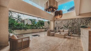A spacious indoor area with a high ceiling, large windows, and a view of greenery outside. There are multiple seating areas with sofas and a wooden bench. - Arbor at Caivama (Sayulita)