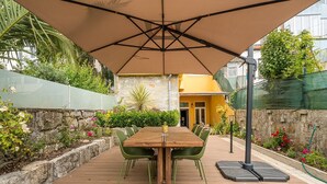 An outdoor patio with a wooden table and chairs, surrounded by plants and a stone wall. - GuestReady - Zenith Building - Room 002 (Porto)