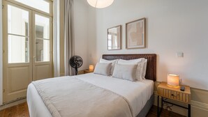 Ein Hotelzimmer mit einem großen Bett, einem Nachttisch mit Lampe, einem Standventilator und zwei gerahmten Bildern an der Wand.
