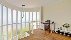 Ein geräumiges Zimmer mit großen Fenstern, Holzboden, einem Schreibtisch mit Stuhl und einem kleinen weißen Regal mit einer Vase.