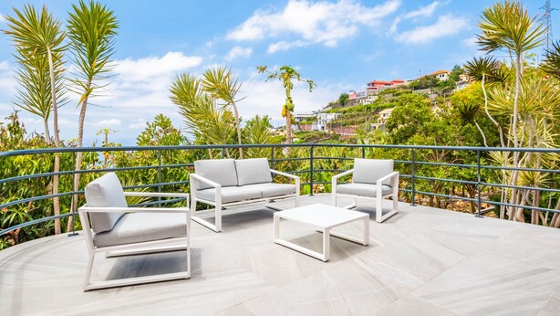 Eine Terrasse im Freien mit weißer Möbel, umgeben von Palmen und Blick auf einen Hügel mit Gebäuden.