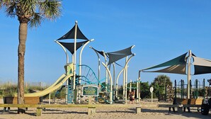 Un parque infantil con toboganes y estructuras para escalar, una palmera y un cartel que indica prohibido estacionar al borde marítimo.