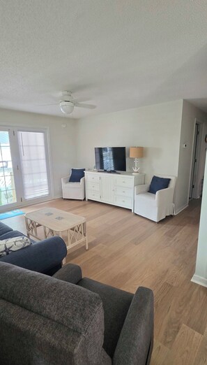 A spacious living room with a flat-screen TV, white furniture, and a ceiling fan.