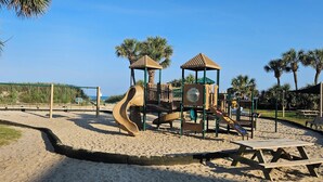 Un parque infantil con toboganes y estructuras para escalar, rodeado de arena y palmeras.