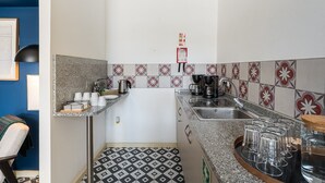 Une cuisine compacte dotée d’un plan de travail en granit, d’une machine à café et d’une bouilloire. On y trouve une petite étagère avec des tasses et un panneau indiquant « GRATUIT ». Le sol est recouvert d’un motif géométrique noir et blanc.