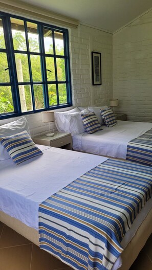 A room with two beds, a window with blue frames, a bedside table with a lamp, and a framed picture on the wall. - Finca La María: piscina infinita, jacuzzi y aves cerca de Armenia (Armenia)