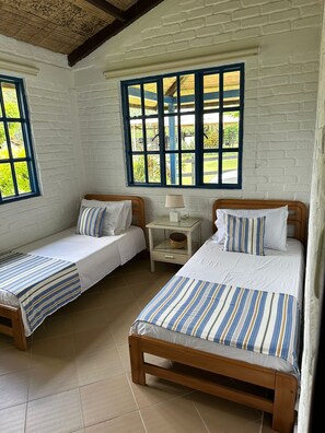A room with two single beds, a small bedside table with a lamp, and a large window with a view of greenery.