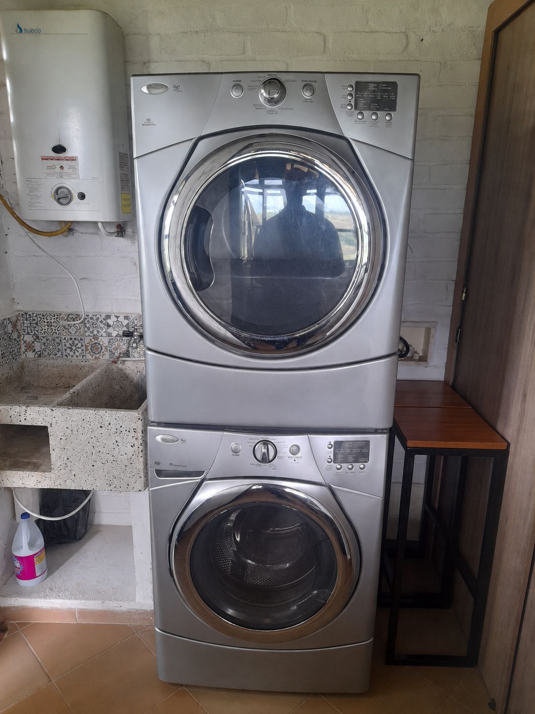 Een gestapelde wasmachine en droger in een wasserij, met een betegelde spatwand en een klein houten tafeltje.