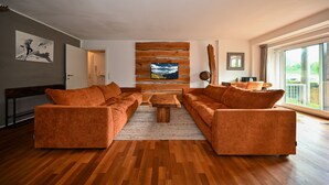 A modern living room with a wooden floor, orange sofas, a coffee table, and a large window with a view.