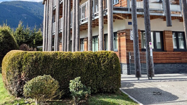 A modern multi-story building with a wooden facade and balconies, surrounded by well-maintained greenery and mountains in the background.